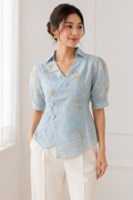 Woman wearing a sky-blue wrap top with ivory floral embroidery, puff sleeves and ivory square buttons.