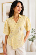 A woman wearing a pale yellow short-sleeve wrap blouse with white floral embroidery and cream pants, standing in a bright indoor room with a framed print and a vase in the background.