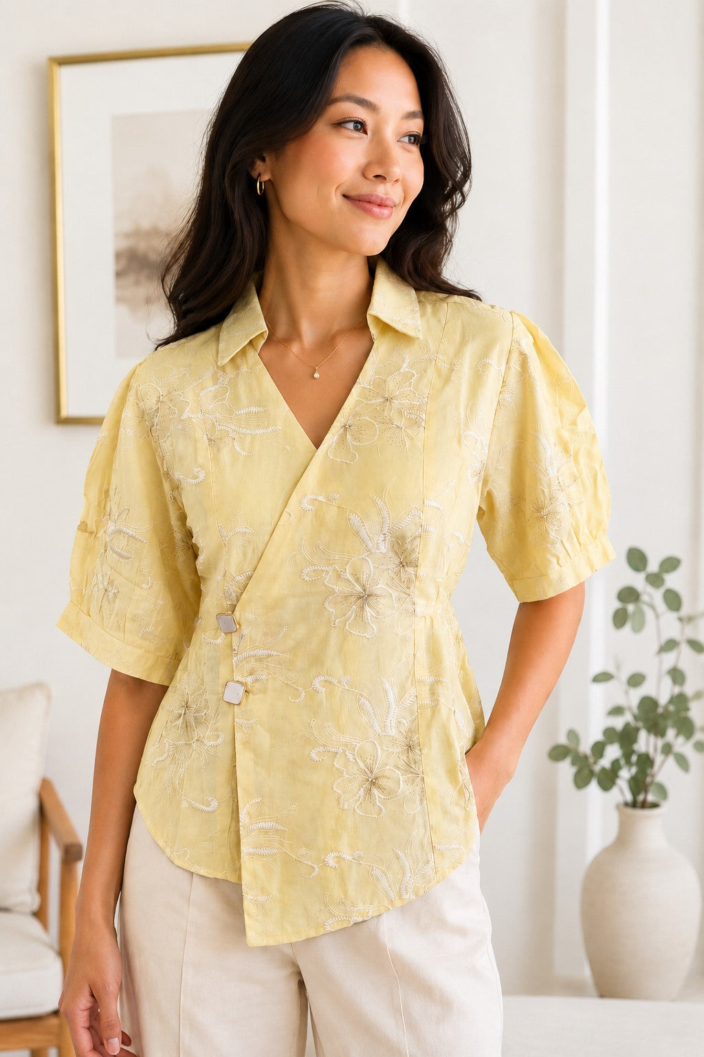 A woman wearing a pale yellow short-sleeve wrap blouse with white floral embroidery and cream pants, standing in a bright indoor room with a framed print and a vase in the background.
